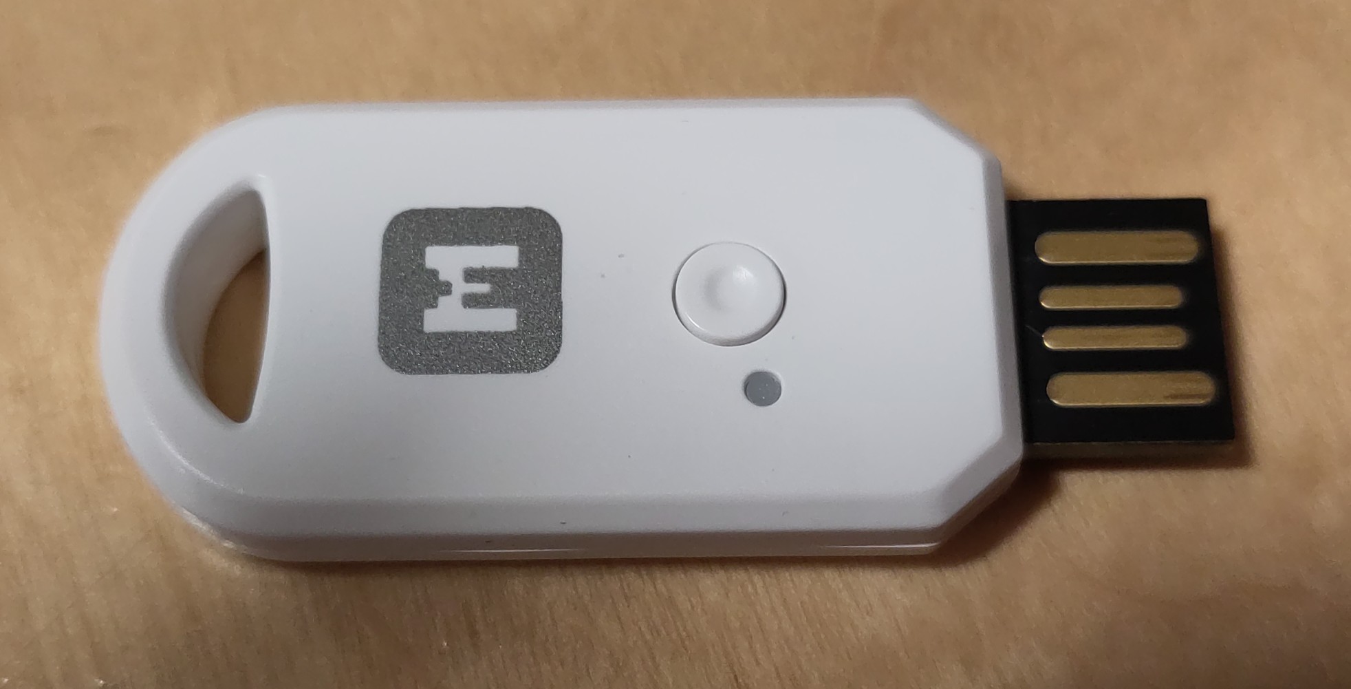 You can see a kind of USB stick lying on a wooden surface. It has an "M" logo printed on it. There is also a small cut-out for a key fob. You can also see a small button and an LED (or that there is a gray dot that can light up). The USB dongle is white.